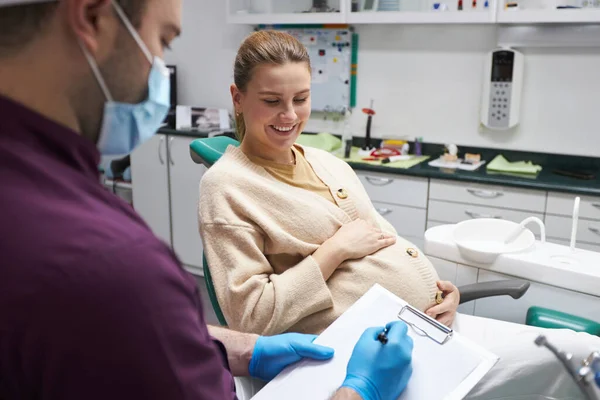 牙医卫生学家给一个年轻的孕妇开了一种牙科预防疗法 坐在牙医的椅子上微笑着 由牙医在怀孕期间进行预防性检查的概念 — 图库照片