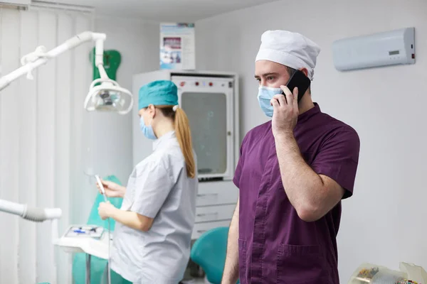 戴着医疗面罩的英俊的正牙医生 一边用手机交谈 一边站在一个现代化的牙科诊所里 背后是在职的怀孕女牙医助理 — 图库照片