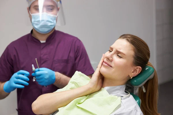 Female patient touching cheek, feeling toothache, waiting for an anesthetic injection for a painless tooth treatment in dental clinic. Doctor dentist in the background holding syringe with anesthetic