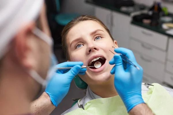 Close-up of young woman with open mouth at dental check-up. Doctor dentist using dental mirror and sterile stainless steel probe, examining teeth and gum of female patient. Dental practice. Dentistry