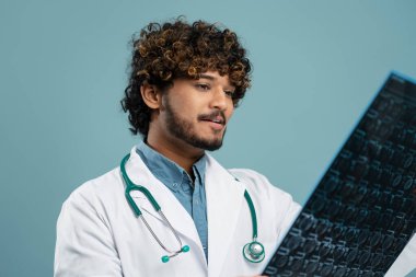 Close-up view of male Indian doctor radiologist or student intern, in white medical gown and stethoscope, analyzing X-ray image on isolated blue background. Healthcare, medicine and diagnosis concept