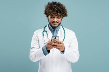 Young Indian man, a doctor intern or student medic in white medical coat with stethoscope, using mobile phone, checks social media content, online consulting patients, isolated over blue background