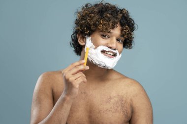 Attractive shirtless Hindu man using razor and shaving foam, shaves, smiles looking at camera, expressing satisfaction by his appearance, isolated on blue background. Copy ad space. Skin care concept