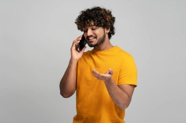 Waist-up portrait of a handsome young Asian bearded man in yellow t-shirt, gesturing while talking on mobile phone, isolated over gray background. People, technology, communication concept
