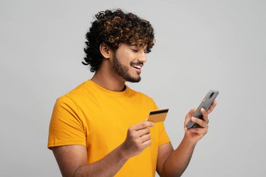 Yakışıklı gülümseyen Asyalı etnik sakallı adam kredi kartı tutuyor ve cep telefonu kullanıyor, çevrimiçi işlemler yaparken veri ekliyor, internet bankacılığı yoluyla para aktarıyor, izole edilmiş gri arka plan
