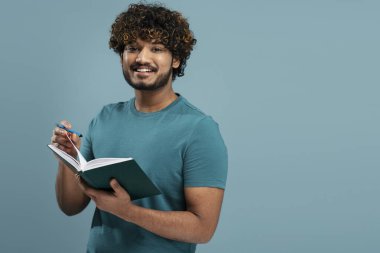 Handsome confident young Asian business man, entrepreneur, student in casual t-shirt, taking notes on his notepad, smiling a toothy smile looking at camera, isolated over blue color background