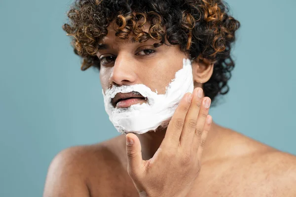 Close-up male beauty portrait of a young shirtless curly-haired Indian man, touching his beard while applying shaving foam on his face on isolated blue background. Skin and body care concept