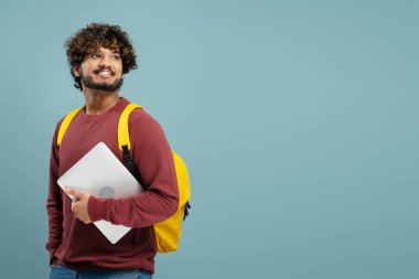 Young Asian man, smiling student in casual clothes, yellow backpack holding laptop PC, isolated on blue wall background. Concept of education in high school, university or college. Mock up. Copy space