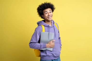 Smart smiling African American student holding laptop computer isolated on yellow background. Education concept. Portrait of happy successful freelancer wearing stylish purple hoodie looking at camera