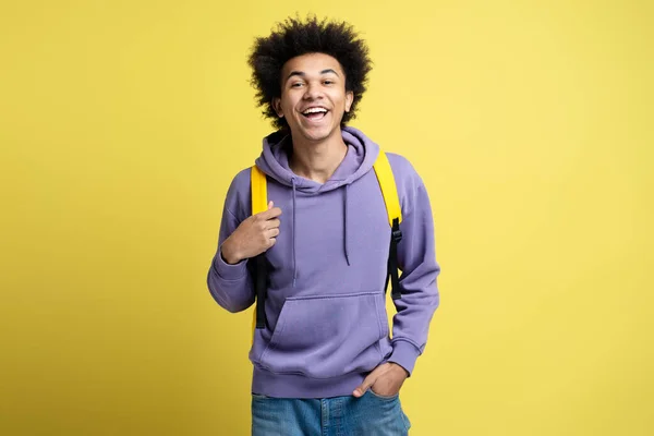 Smart smiling university student with backpack looking at camera isolated on yellow background. Education concept