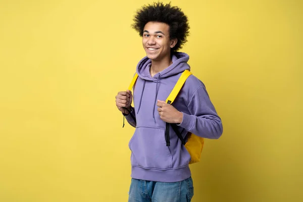Attractive smart smiling university student with backpack looking at camera isolated on yellow background, copy space. Education concept