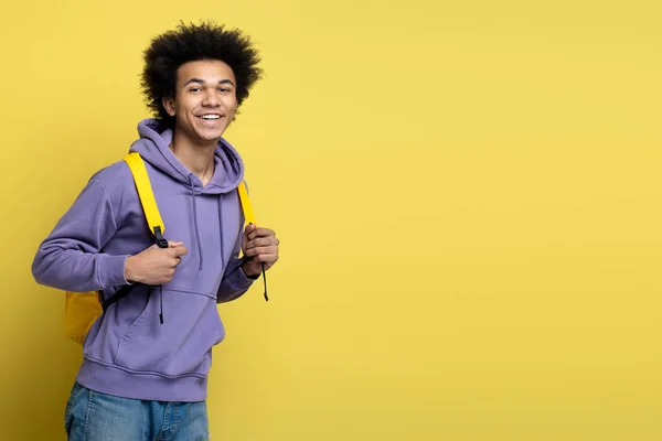 Portrait of young smiling university student with backpack wearing stylish hoodie looking at camera isolated on yellow background, copy space. Education concept