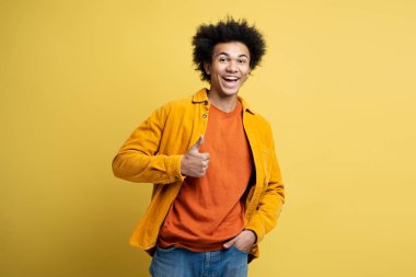 Young emotional African American man wearing stylish colorful outfit holding thumb up looking at camera isolated on yellow background
