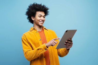  Smiling African American man holding digital tablet watching videos isolated on blue background. Happy attractive male with curly hair using mobile app, ordering food, shopping online. Technology