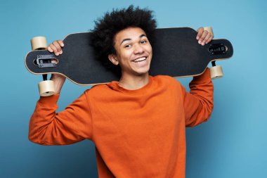 Young smiling African American man holding longboard isolated on blue background. Portrait of attractive happy skater looking at camera. Street culture, active lifestyle concept