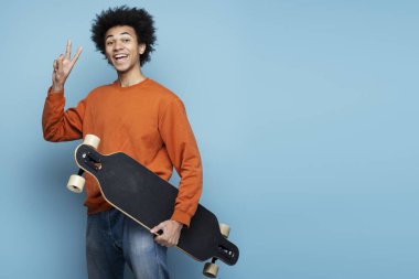 Handsome smiling African American man holding longboard showing victory sign isolated on blue background. Portrait of attractive happy skater looking at camera, copy space  