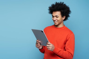 Confident smiling African American man using digital tablet watching videos on website isolated on blue background, copy space. Happy attractive male shopping online with sales. Technology concept