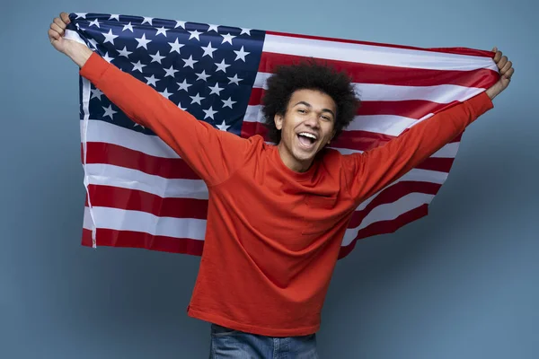 Young happy African American man holding American flag isolated on blue background. Emotional patriot celebration Independence day looking at camera