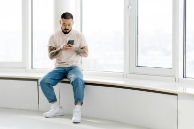Serious handsome Korean man  holding mobile phone shopping online sitting on windowsill in white office. Young hipster guy with stylish tattoos on his hands chatting, reading text message, copy space