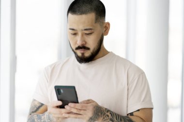 Serious handsome Asian man using mobile phone , shopping online at home. Hipster guy with stylish tattoo chatting, communication online. Technology concept