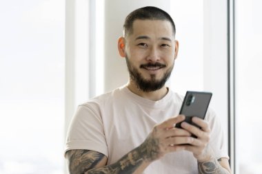  Smiling Asian man with stylish tattoos holding smartphone using mobile app shopping online at home. Modern bearded hipster ordering food. Mobile banking, technology concept