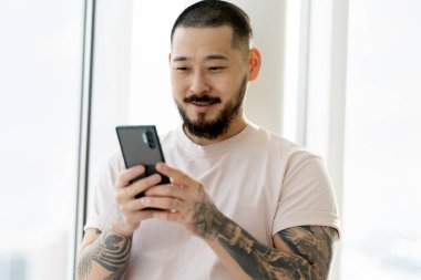 Smiling Asian man with stylish tattoos holding  smartphone using mobile app shopping online at home. Modern bearded hipster reading text message, ordering food, communication. Technology concept