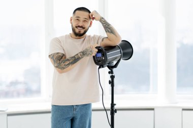 Smiling photographer's assistant supports his head with his hand, leaning on a lamp. Positive asian man with a beard and stylish tattoos on his hands in a modern photo studio, copy space