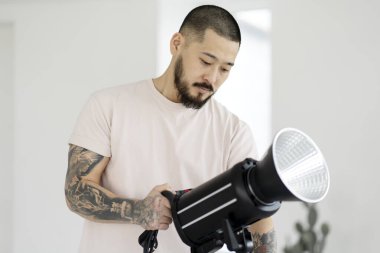 Portrait of professional asian photographer with stylish tattoos setting up lighting in modern studio. Assistant preparing for photo shoot