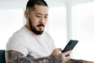 Serious handsome Asian man holding smartphone, checking mail, shopping online at home. Bearded hipster guy with stylish tattoos using mobile app, watching videos