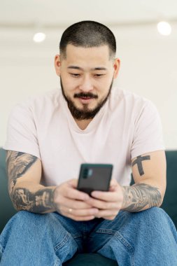 Young smiling bearded man using mobile phone, searching on website, indoors. Handsome hipster with modern tattoos on his hands holding smartphone, shopping online, ordering food sitting at home 