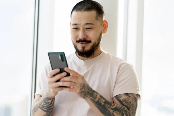 Smiling Asian Man Stylish Tattoos Holding Smartphone Using Mobile App — Stockfoto