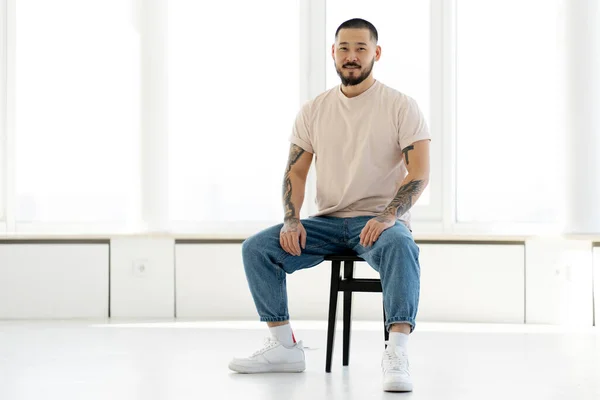 Handsome korean man with stylish tattoos sitting in a casual style of clothes on a chair in a white office against the background of windows. Asian model posing for pictures