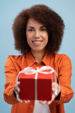 Satisfied young female holding out red wrapped gift box, giving present, looking smiling at camera, wearing casual style clothes. Indoor studio shot isolated on blue background 