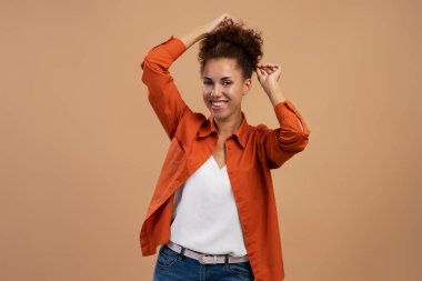 Portrait of cheerful curly african woman preparing hairstyle and looking at camera with toothy smile facial expression. Indoor studio shot isolated on brown background 