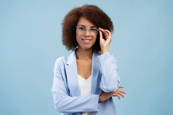 笑着非洲裔美国女企业家站在那里 看着被蓝色背景隔离的相机 快乐的女商人 戴着时髦的眼镜 手挽手交叉着 — 图库照片