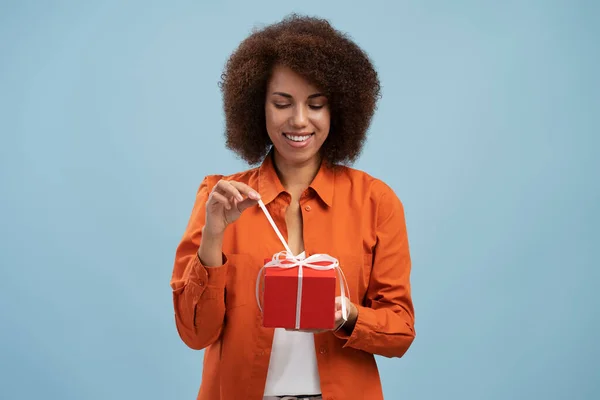 Portrait of african american woman opening gift box, unwrapping birthday present for celebrating holiday, pulling ribbon, looking at gift. Indoor studio shot isolated on blue background