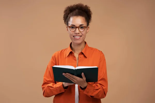 アフリカ系アメリカ人女性が茶色の背景に孤立した本を読んで笑顔の肖像画 カメラを見て勉強する学生 教育の概念 — ストック写真