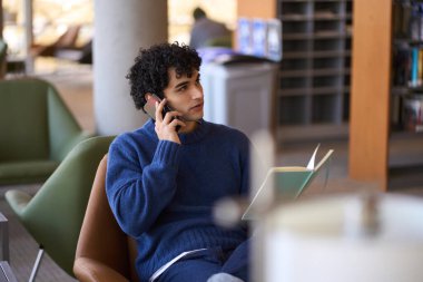 Genç Latin Amerikalı iş adamı, serbest girişimci, cep telefonuyla konuşan, elinde defteriyle kütüphanede sandalyede oturan. Halk, iş, uzaktan çalışma ve eğitim kavramı