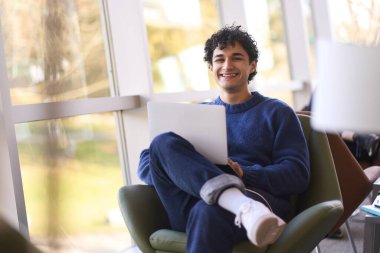 Mavi süveter ve kot pantolon giymiş yakışıklı, neşeli dişlek bir gülümsemeyle kameraya bakan, modern ofis içinde dizüstü bilgisayarla çalışan yakışıklı bir İspanyol. Uzaktan kumanda. İnternet işi. İnsanlar