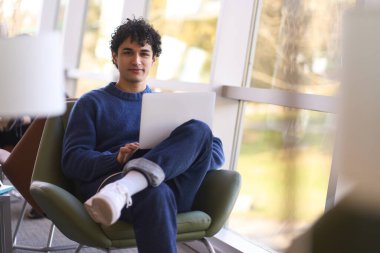 Genç, başarılı, Latin Amerikalı serbest iş adamı bir erkeğin portresi. Günlük giysiler içinde, büyük pencerelerin yanındaki koltukta oturuyor, dizüstü bilgisayarı elinde, kameraya bakıyor..