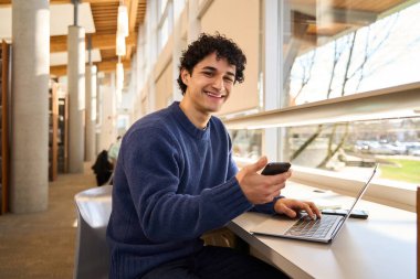 Cep telefonlu zeki bir öğrenci, kameraya bakıp gülümsüyor, kütüphanede dizüstü bilgisayarla oturuyor.