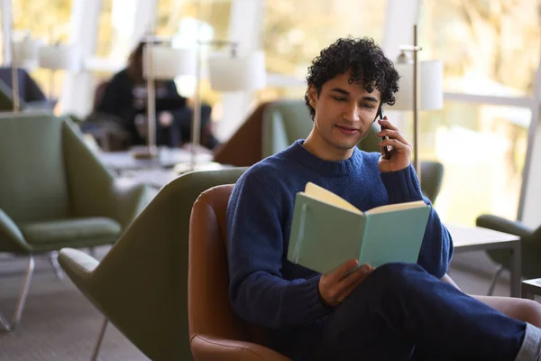 本を読んでいる間に携帯電話で話しているリラックスした若いヒスパニック系の男性は 椅子に座っています 成功した起業家のフリーランスが会議を計画していると確信しています彼のノートブックで彼のノートを見て — ストック写真