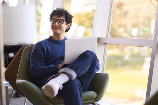 Confident Hispanic guy, smart university student, in eyeglasses, smiling and looking aside, browsing websites, surfing on internet, preparing exams and working onine on diplome project, using laptop