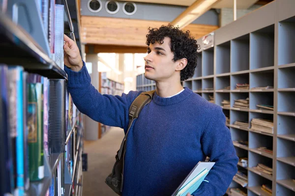 図書館のキャンパスの本棚から本を選び 大学院生のバックパック付きのカジュアルな服を着たハンサムな若い暗い髪の巻きラテンアメリカ人男性 人々と教育の概念 — ストック写真