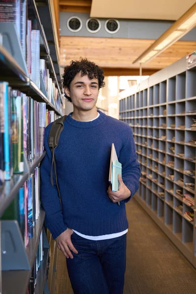 若いラテン系アメリカ人の男性学生がバックパックを持っていたり 教科書やノートパソコンを手に持っていたり 図書館のキャンパスや書店の本棚のそばに立っていたり カメラを見て笑顔を浮かべています 教育を受けた人々の概念 — ストック写真
