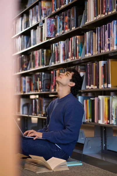 ラテン系アメリカ人の学生がノートパソコンを使って勉強し 大学図書館のキャンパス内に文学や本が置かれた棚の上に座っている 技術だ — ストック写真