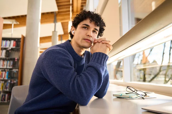 若いラテンアメリカの男性学生は イヤホンを使用してオンライン講義を聞き ノートパソコンで机に座って 現代の革新的な大学図書館キャンパスで勉強します 教育理念 — ストック写真