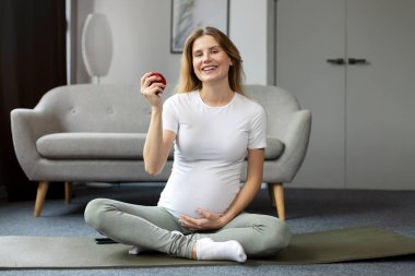 Genç, gülümseyen, yoga yapan, Lotus pozu veren, elinde kırmızı elma tutan hamile bir kadın. Hamilelik yogası, sağlıklı gıda konsepti