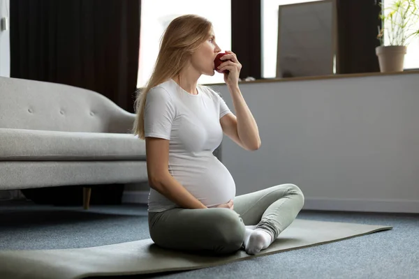 妊娠中の若い女性は 自宅でハスのポーズに座ってヨガを練習赤いリンゴを食べている 妊娠中のヨガ 健康食品の概念 — ストック写真