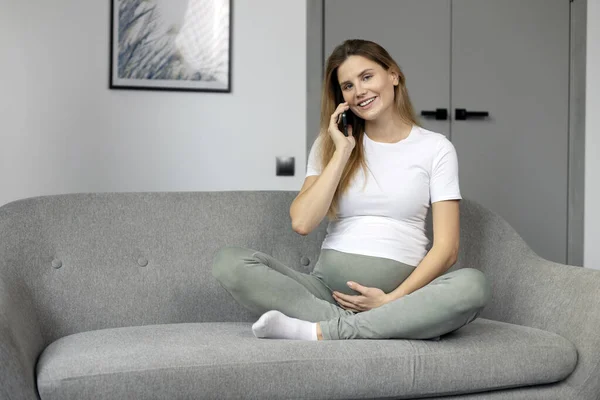 若い笑みを浮かべて妊娠中の女性の携帯電話のソファの上に自宅で座って話している 技術コンセプト — ストック写真